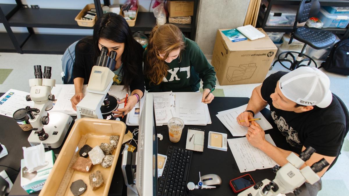 two students take notes while another looks at rock samples under a microscope